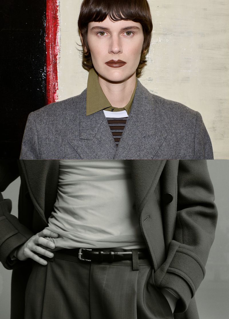 Fashion portrait of a model wearing a grey jacket and pants, hands on hips, with a painted background, styled with brown lipstick and a blunt hairstyle.