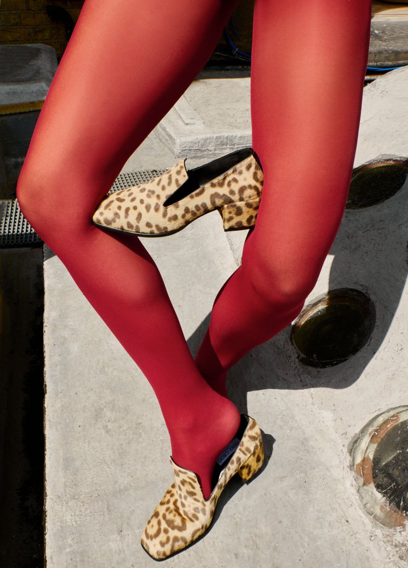 Close-up of woman's legs in red tights with animal-print heeled loafers, posing on a grey concrete surface in natural lighting.