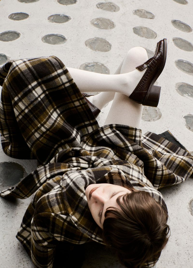 Woman lies on a patterned concrete surface wearing a plaid skirt, tights, and brown heeled shoes showcasing a vintage aesthetic.