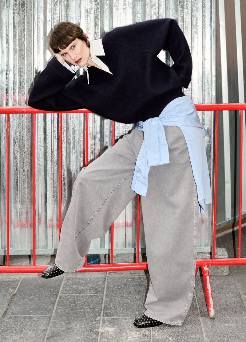 Fashionable adult woman in a dark sweater and wide leg pants leaning against a red barricade on a city street looking at the camera.
