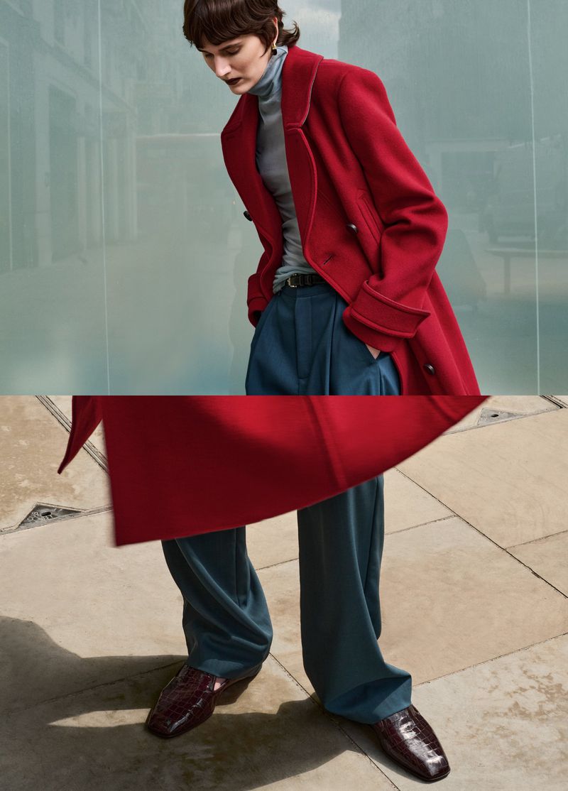 Model wearing a red coat and blue trousers standing on paved outdoor ground