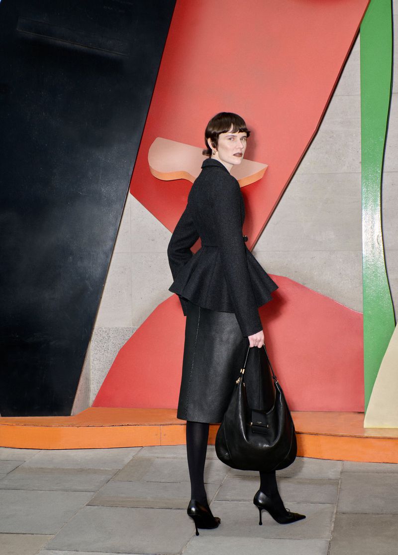 A stylish adult woman in a black peplum jacket, pencil skirt, and heels holds a black handbag against a red and black geometric wall.