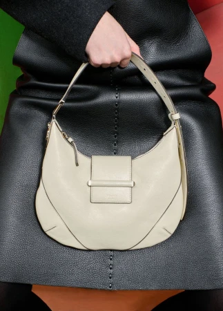 Close-up of a person holding a cream-colored leather handbag against a black textured garment.