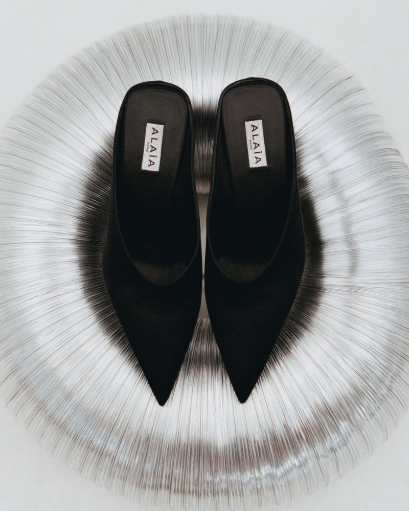 Elevated view of a pair of black Alaïa heeled mules centered on a reflective metallic silver, textured round surface and a bright white background.