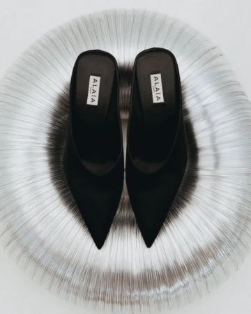 Elevated view of a pair of black Alaïa heeled mules centered on a reflective metallic silver, textured round surface and a bright white background.