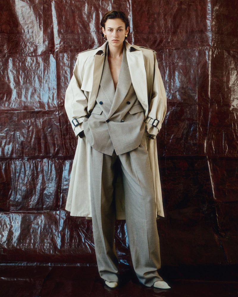 A woman in a beige suit and trench coat poses against a textured brown background, showcasing a blend of classic and modern fashion.