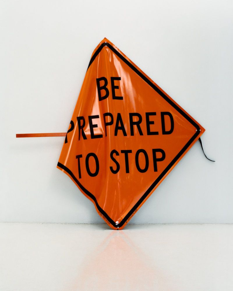 An orange, reflective temporary traffic sign reads 'Be Prepared to Stop' against a stark white background in a studio setting.