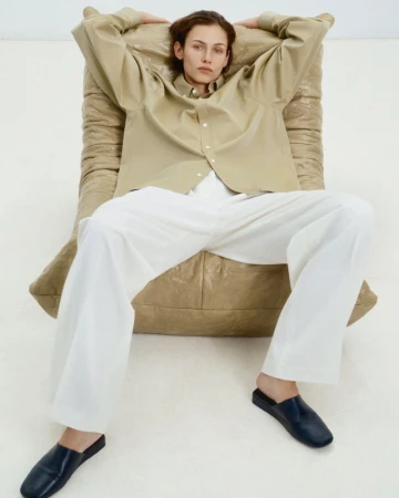 A relaxed adult model in a tan button-down shirt and white pants reclines on a padded chair in a bright studio with black leather mules.