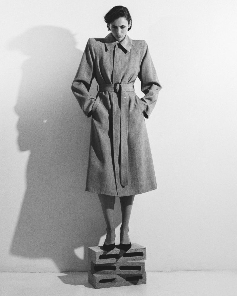 Monochrome full shot of a fashion model in a belted overcoat posing on stacked concrete blocks in a studio setting with a wall shadow.