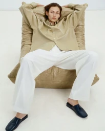 Model seated on a quilted beige chair wearing an oversized tan shirt and white trousers against a white background