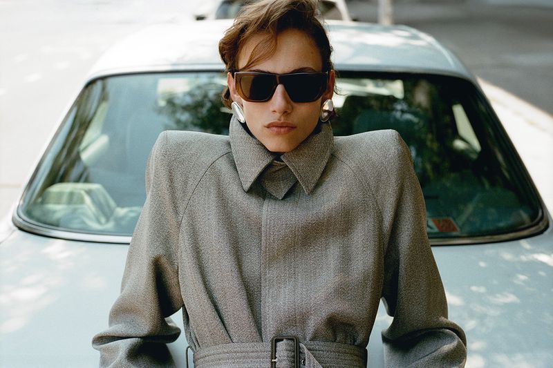 Model leaning on a car wearing a structured grey coat and sunglasses with large earrings