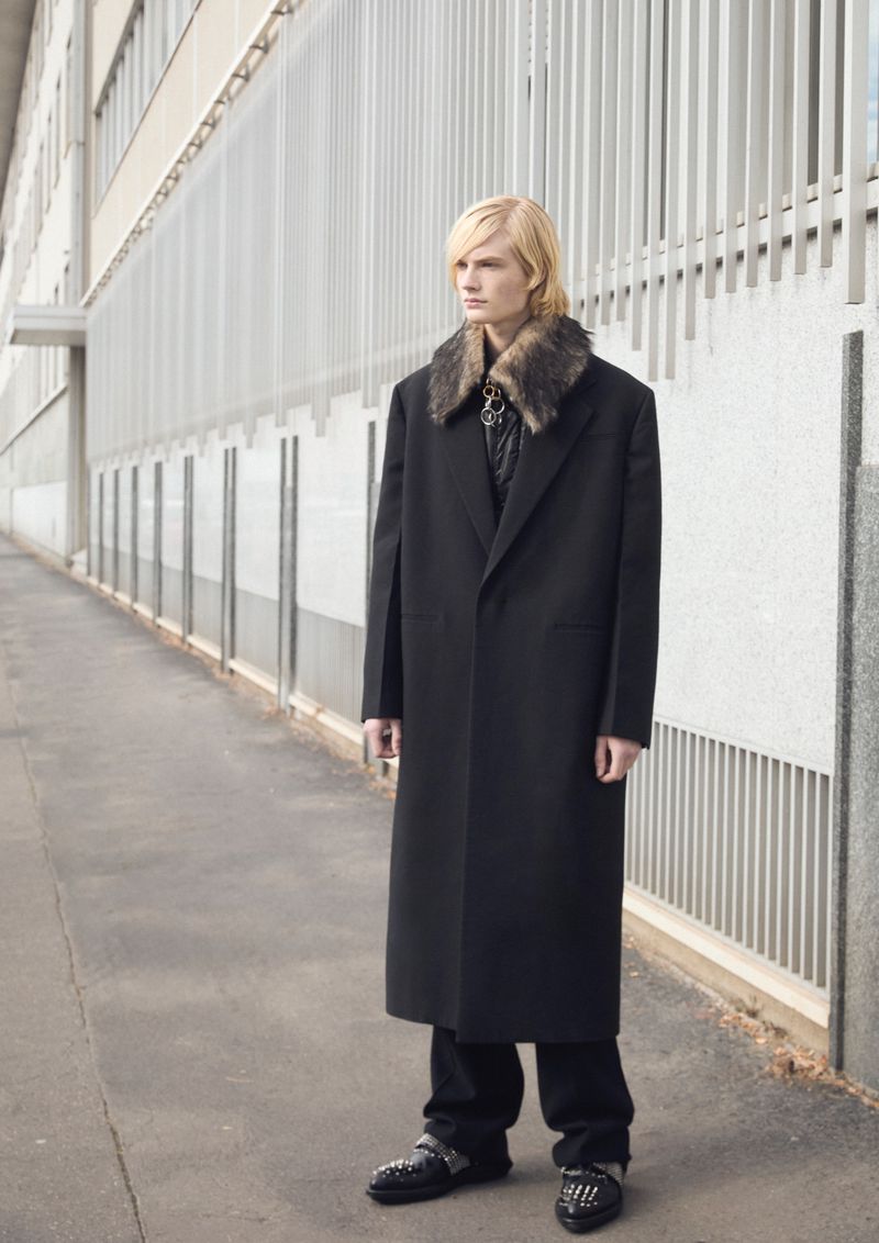 Fashionable male model with blond hair poses in a long black coat with a fur collar outside of a concrete building on a sunny day.