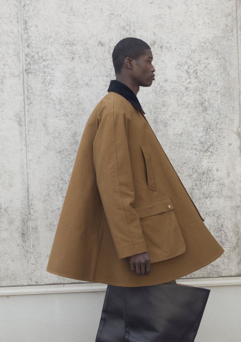 Model wearing a brown A-line jacket with dark collar and holding a black tote bag against a concrete wall