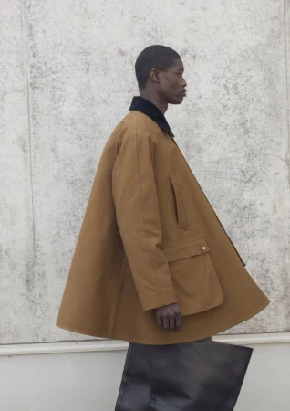 Model wearing a brown A-line jacket with dark collar and holding a black tote bag against a concrete wall