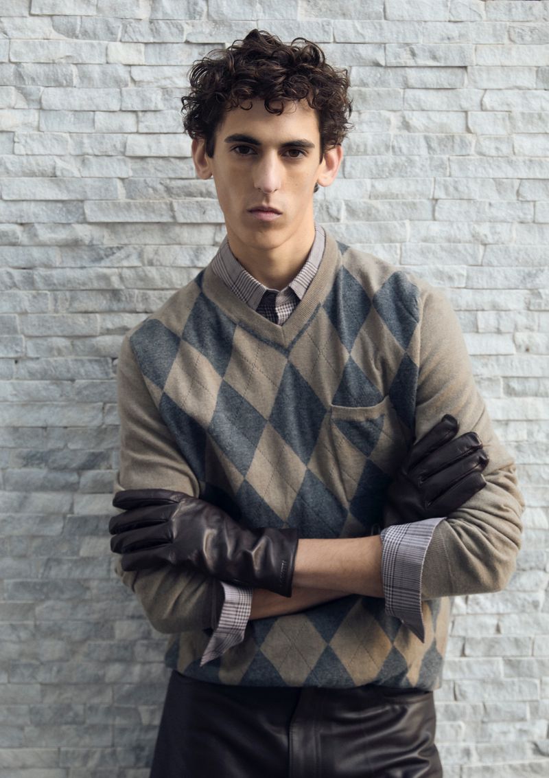 A young adult man with curly hair poses indoors wearing a patterned argyle sweater, buttoned shirt, black leather gloves, and leather pants.