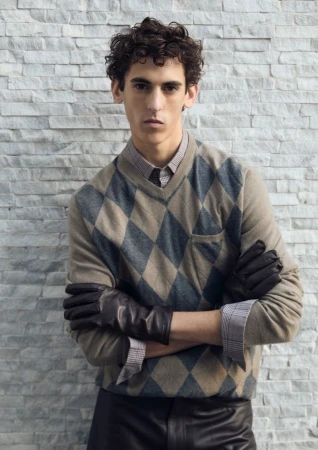 A young adult man with curly hair poses indoors wearing a patterned argyle sweater, buttoned shirt, black leather gloves, and leather pants.