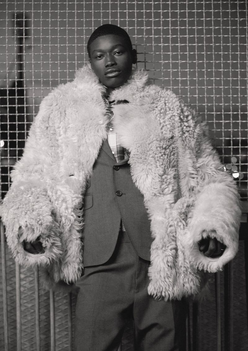 Portrait of a Black man wearing a large fur coat over a suit, posing in front of a metal grid wall in a grayscale image.