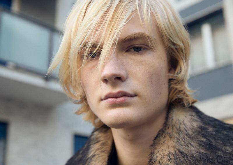Portrait of a fair-haired young man with freckled skin wearing a fur-trimmed jacket against an urban building backdrop.