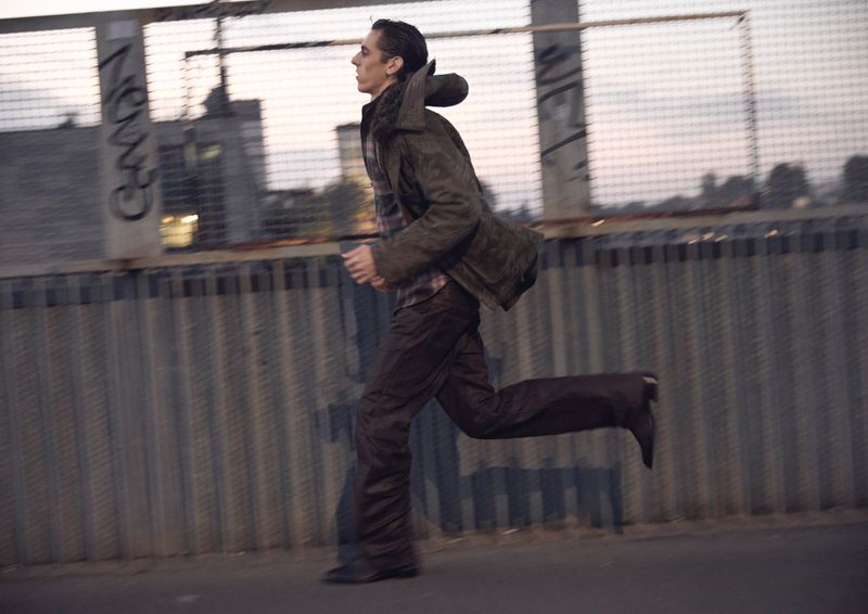 A man running outdoors on a bridge, wearing a coat and trousers, captured in motion with blurred background, and side view.