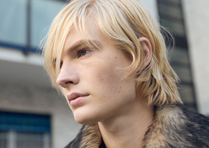 Close-up of a blonde man with light skin wearing a grey and black fur-collared jacket in an outdoor setting with a white building.