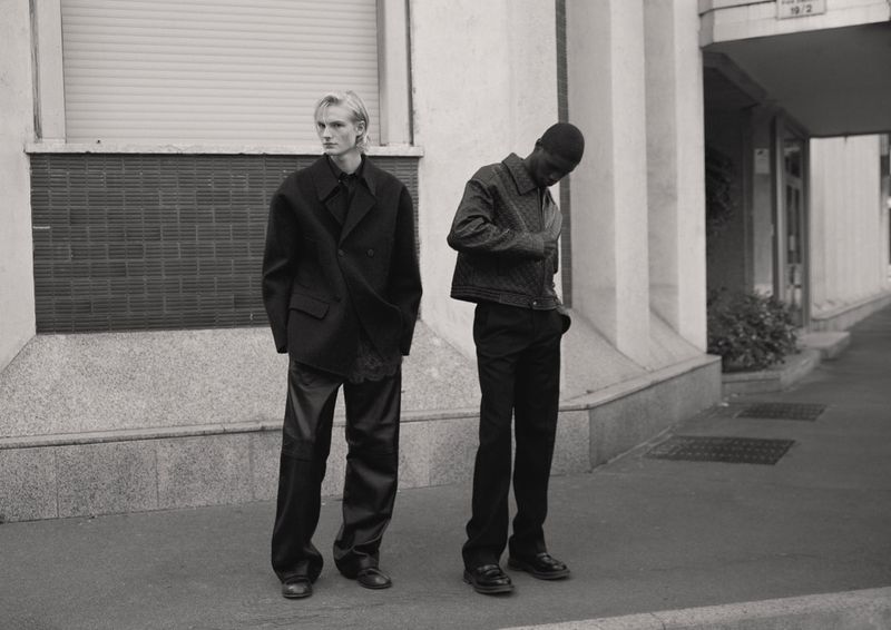 Two fashionably dressed men pose outdoors in monochrome: one in a blazer and the other in a jacket, captured in medium shot.