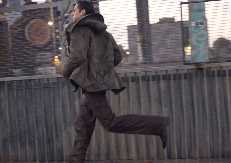 Model wearing a dark hooded jacket and matching trousers running along a metal bridge with urban structures in the background.