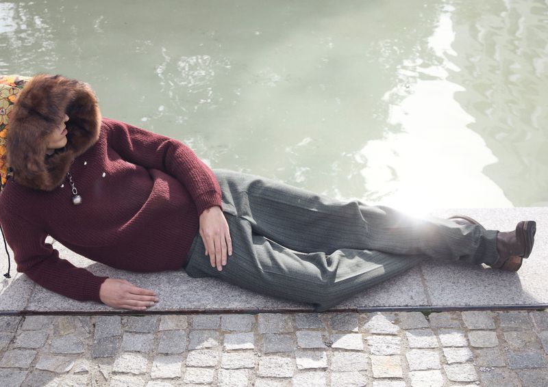 Woman in maroon sweater and striped pants lying on stone next to water on a sunny day. Fur hood frames her face in a relaxed pose.