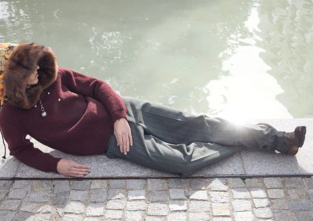 Woman in maroon sweater and striped pants lying on stone next to water on a sunny day. Fur hood frames her face in a relaxed pose.