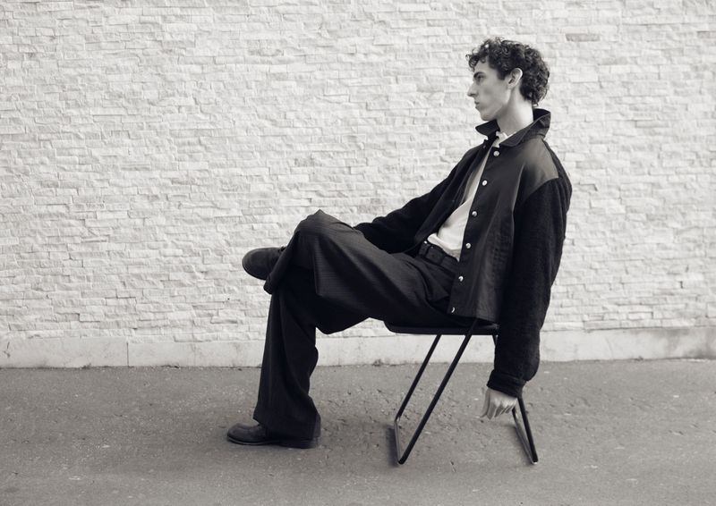 Profile of a seated man wearing a dark jacket and trousers on a metal chair against a textured stone wall.