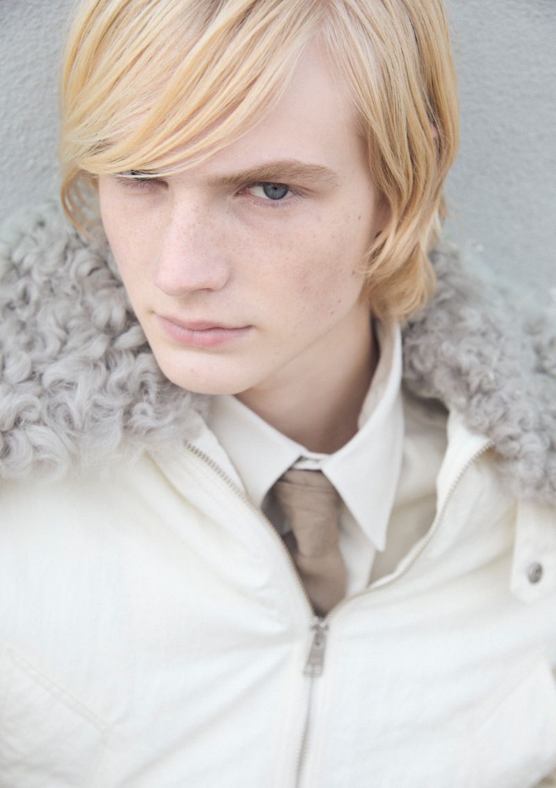 Close-up of a blond adult male with blue eyes wearing a white jacket, shirt, and tie, against a textured gray background in portrait orientation.