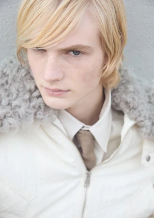 Close-up of a blond adult male with blue eyes wearing a white jacket, shirt, and tie, against a textured gray background in portrait orientation.