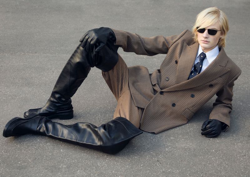 Fashion photo of a blonde adult male model in a tan suit, white shirt, dark tie, black gloves and boots, laying on grey pavement.
