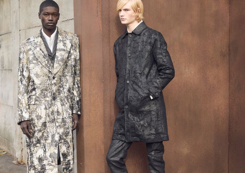 Two men in patterned overcoats stand against a textured wall in an outdoor fashion shot, capturing a stylish editorial moment.
