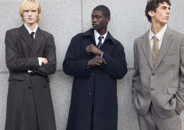 Three diverse men wearing suits and overcoats pose against a concrete wall, showcasing formal fashion in an outdoor setting.