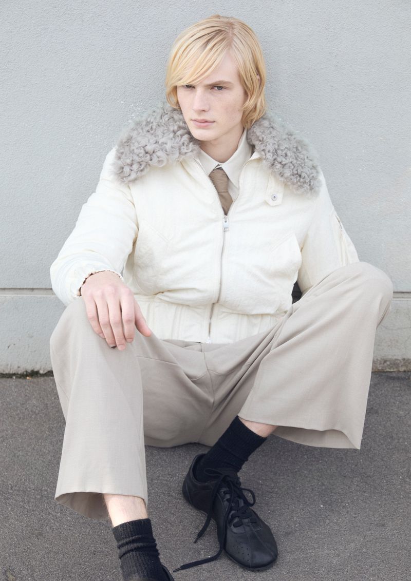 Blonde male model wearing white jacket with fur collar, cream trousers and black sneakers seated outdoors.