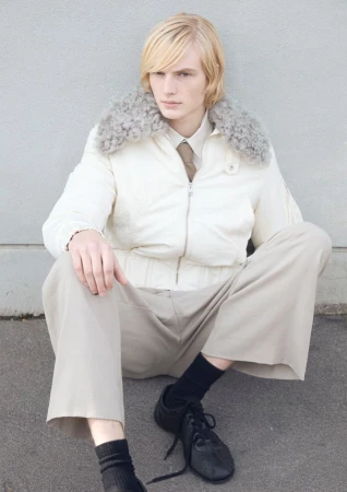 Blonde male model wearing white jacket with fur collar, cream trousers and black sneakers seated outdoors.