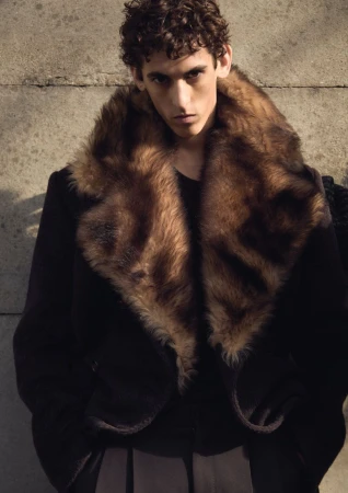 Model with curly hair wearing a dark coat with a large fur collar against a stone wall
