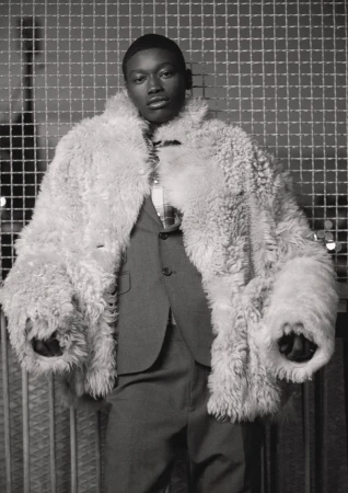 Black man wearing a large fluffy shearling coat and tailored suit standing against a wire mesh screen.