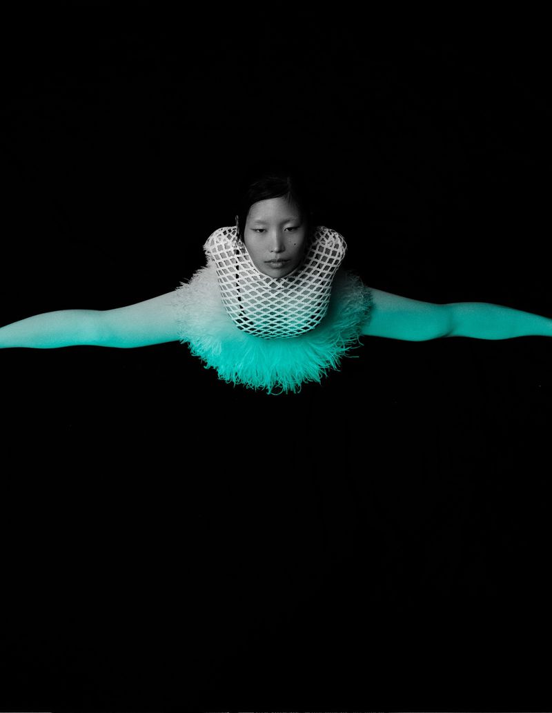 Asian woman posing in a white neck piece and blue-green bodysuit against a black backdrop, arms stretched out, fashion forward, studio shot.