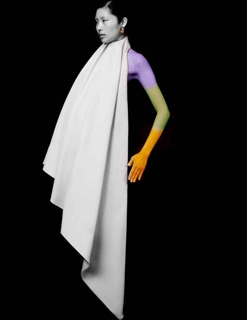 Asian woman in a draped white garment, digitally manipulated with purple, green, and orange sections on her arm, set against a black backdrop.