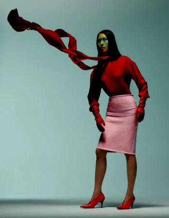 Woman in red blouse and pink skirt poses with a red scarf against a light blue backdrop in a studio fashion shot.