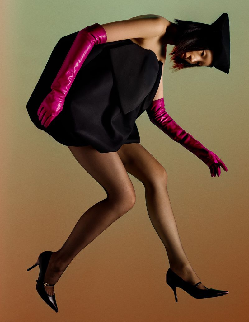Woman poses in a black dress with bright pink gloves and black heels against a blended background of tan, green, and brown.