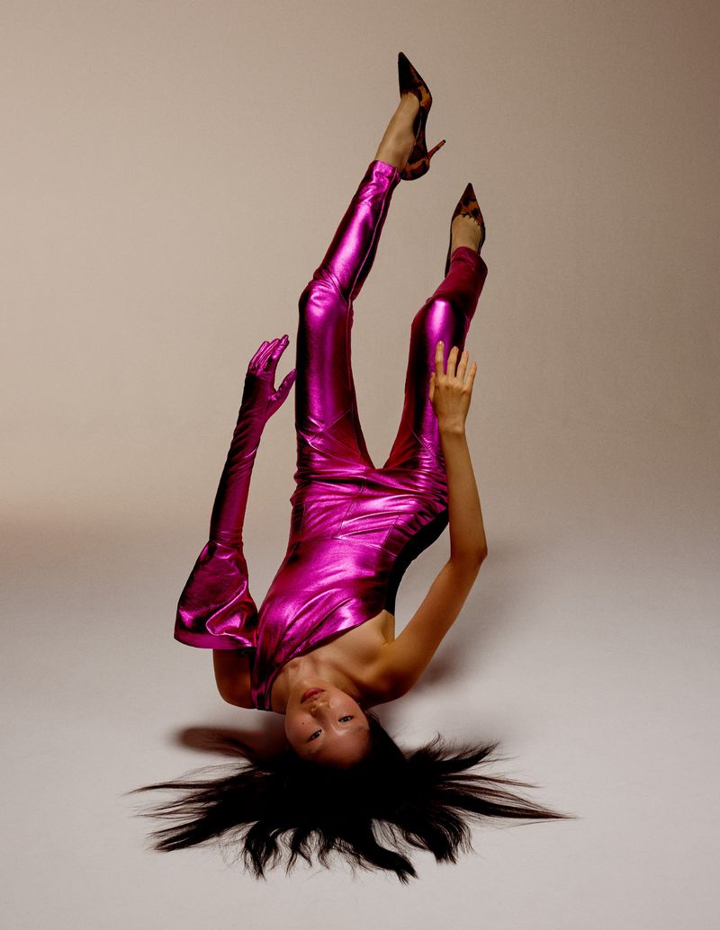 A fashion shot features an Asian woman in a metallic pink jumpsuit and heels, lying upside down against a neutral backdrop in a studio setting.