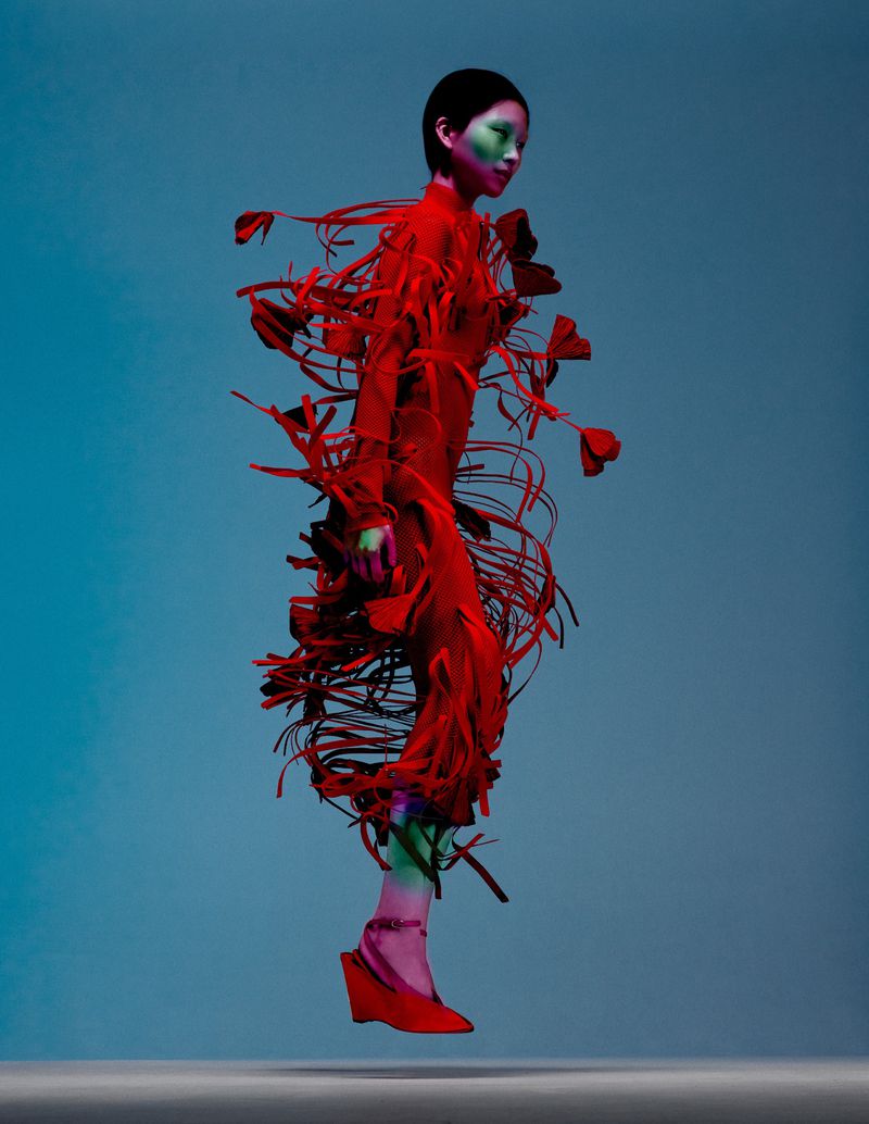Side profile shot of a person wearing a red dress with fringed accents and matching red wedge shoes, posing against a gradient blue background.
