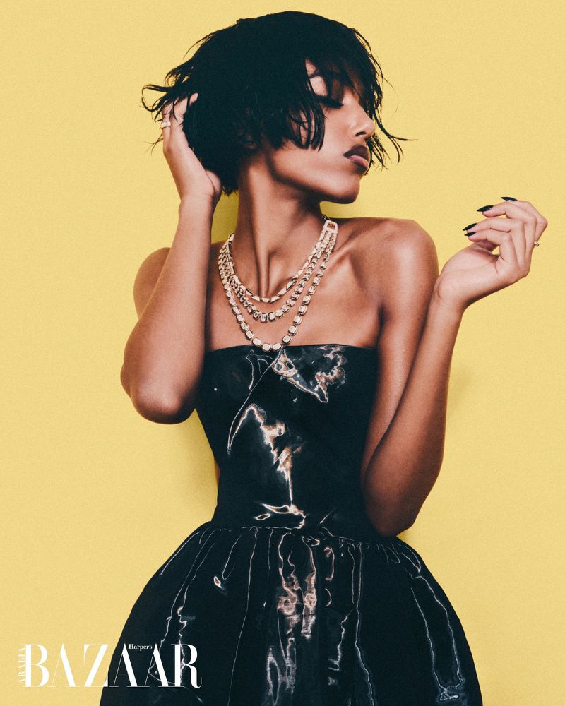 Fashion shot of a Black woman in a black dress and layered pearl necklace against a bright yellow background, looking to the side.