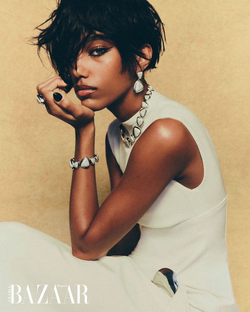 Close-up fashion shot of a Black female model in a white dress and jewelry, resting her face on her hand against a tan backdrop.