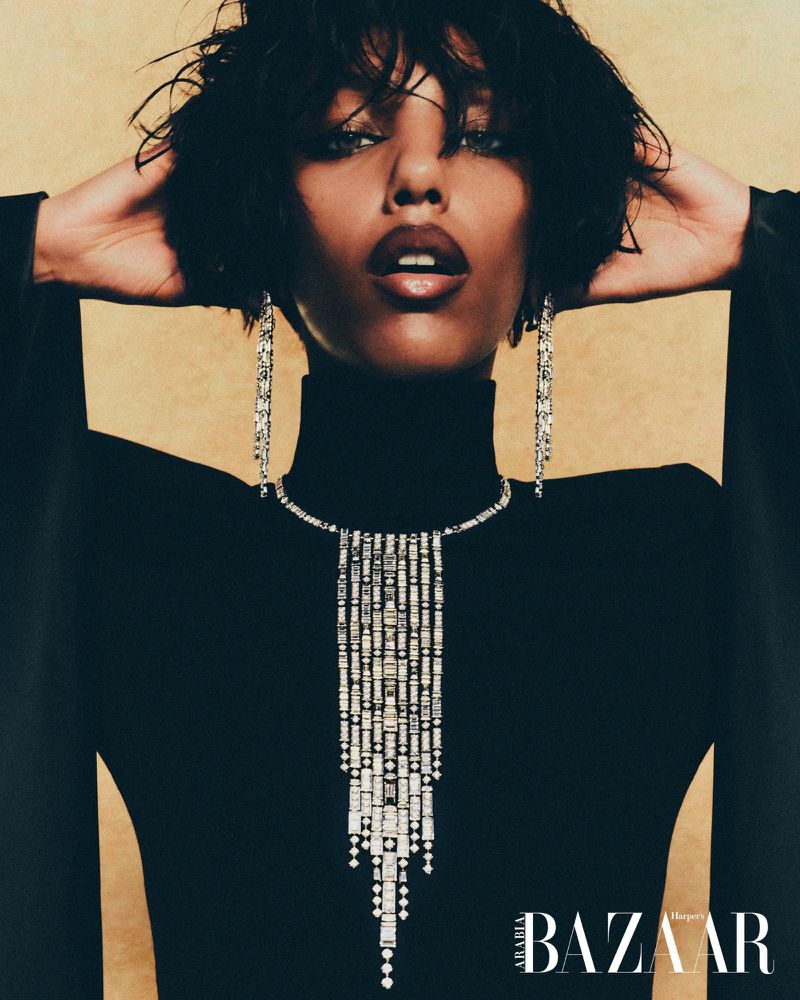 A dark-haired woman poses in a black turtleneck and jewelry against a gold background in a portrait fashion shot.