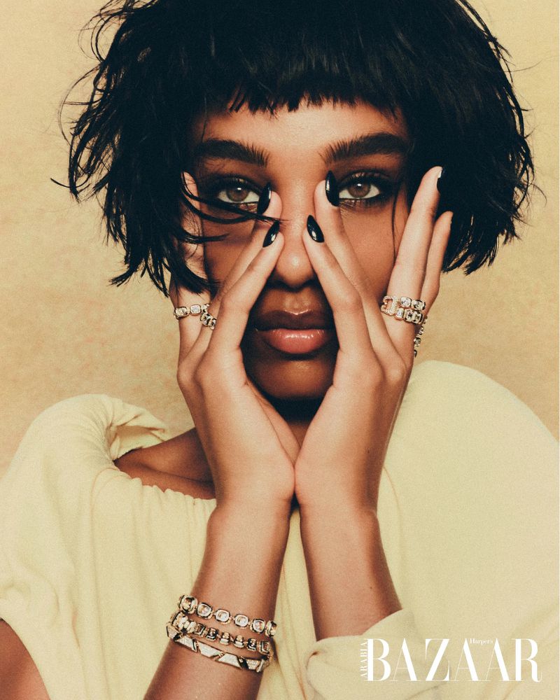Close-up fashion shot of a woman with dark hair posing with her hands near her face, wearing jewelry, and a yellow top indoors.