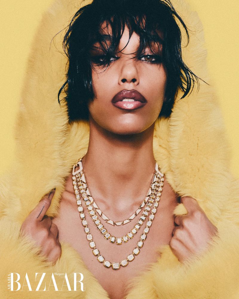 Fashion portrait of a woman wearing a yellow faux fur hooded coat and a layered gold necklace against a yellow background.