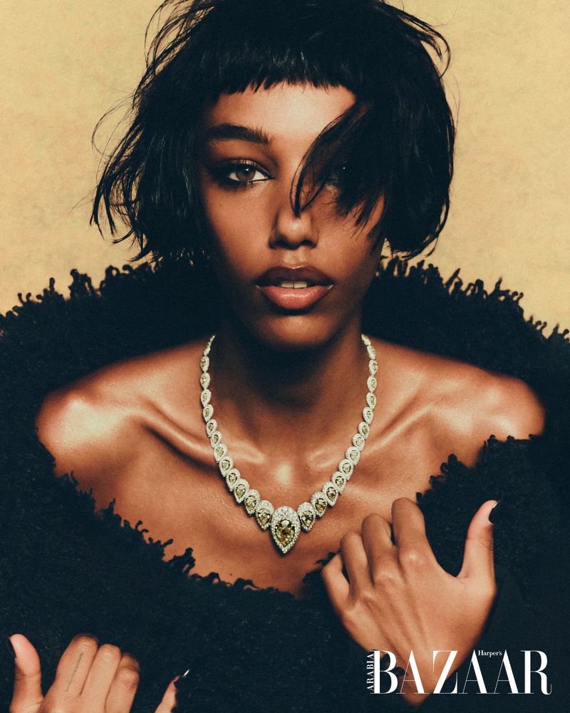Close-up portrait of an adult woman wearing a jewel-encrusted necklace, a textured black jacket, and natural makeup against a tan background.